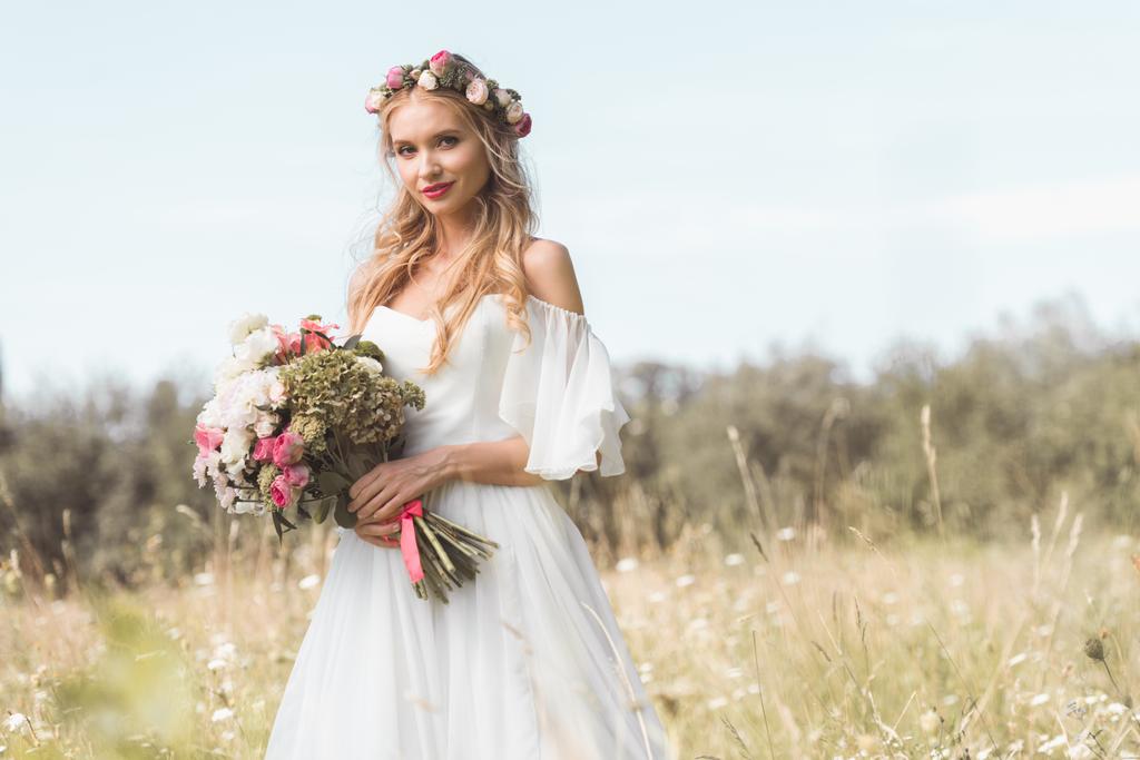beautiful young bride with wedding bouquet standing on blooming field and smiling at camera - Photo, Image