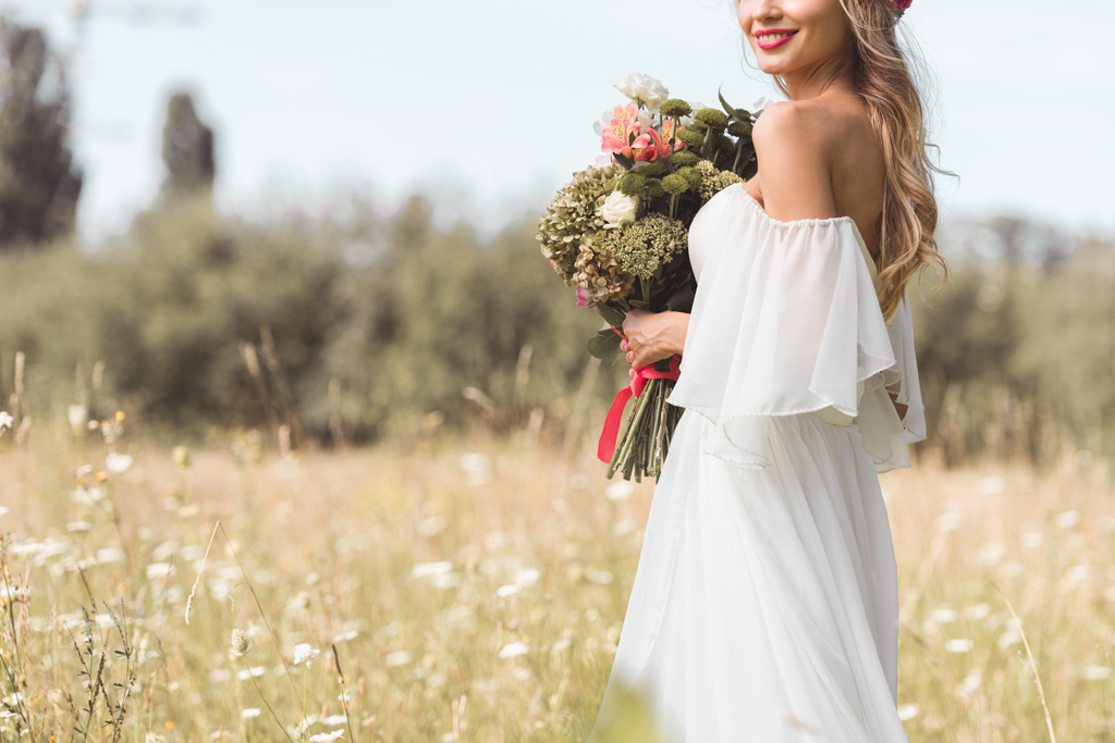 обрезанный снимок улыбающейся молодой невесты в белом платье, держащей свадебный букет на открытом воздухе
 - Фото, изображение