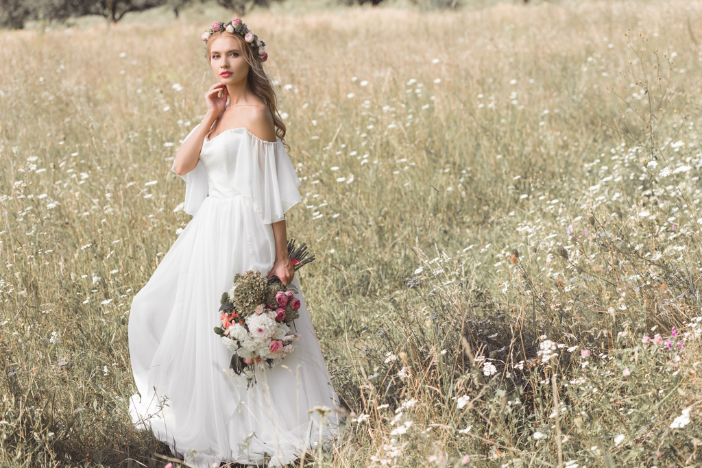 krásná zamyšlená mladá nevěsta ve svatebních šatech a květinový věnec drží kytici květin stoje venku - Fotografie, Obrázek