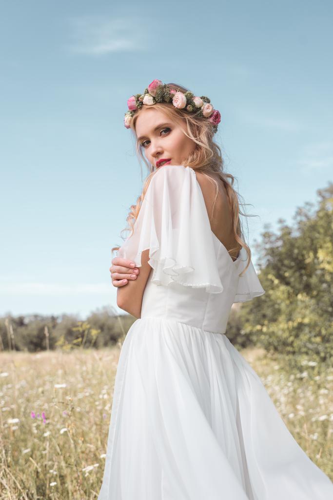 ніжна молода наречена у весільній сукні та квітковому вінку, що стоїть на полі і дивиться на камеру
 - Фото, зображення