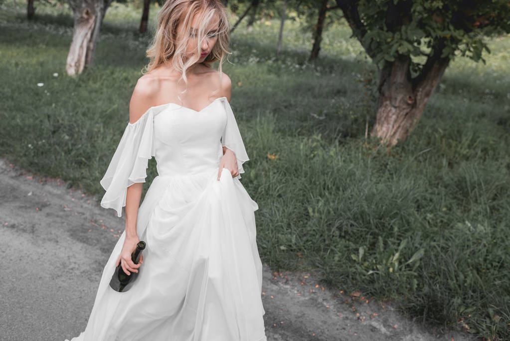 Blick aus der Vogelperspektive auf eine aufgebrachte junge Braut im Brautkleid, die eine Flasche Wein in der Hand hält und im Park spaziert  - Foto, Bild