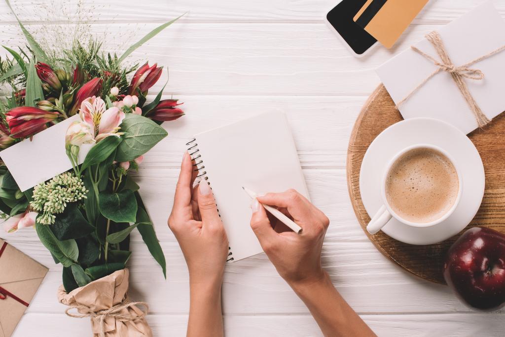 vista parcial de la mujer haciendo notas en el cuaderno en la mesa con ramo de flores y taza de café
 - Foto, Imagen
