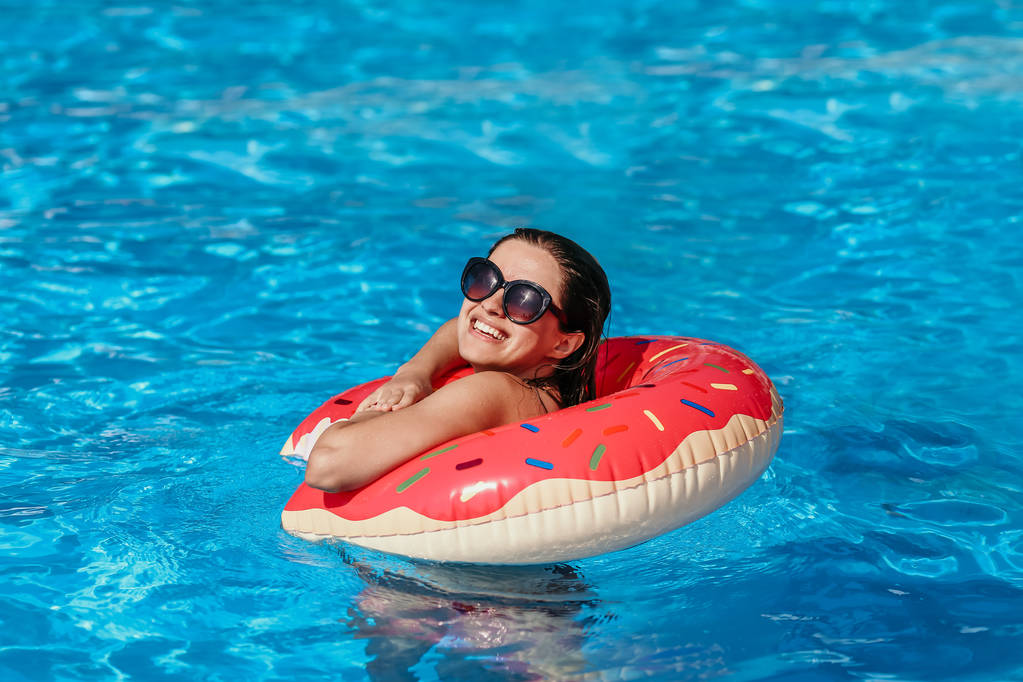 Mooie jonge vrouw met roze donut cirkel hebben rest in blauwe zwembad - Foto, afbeelding