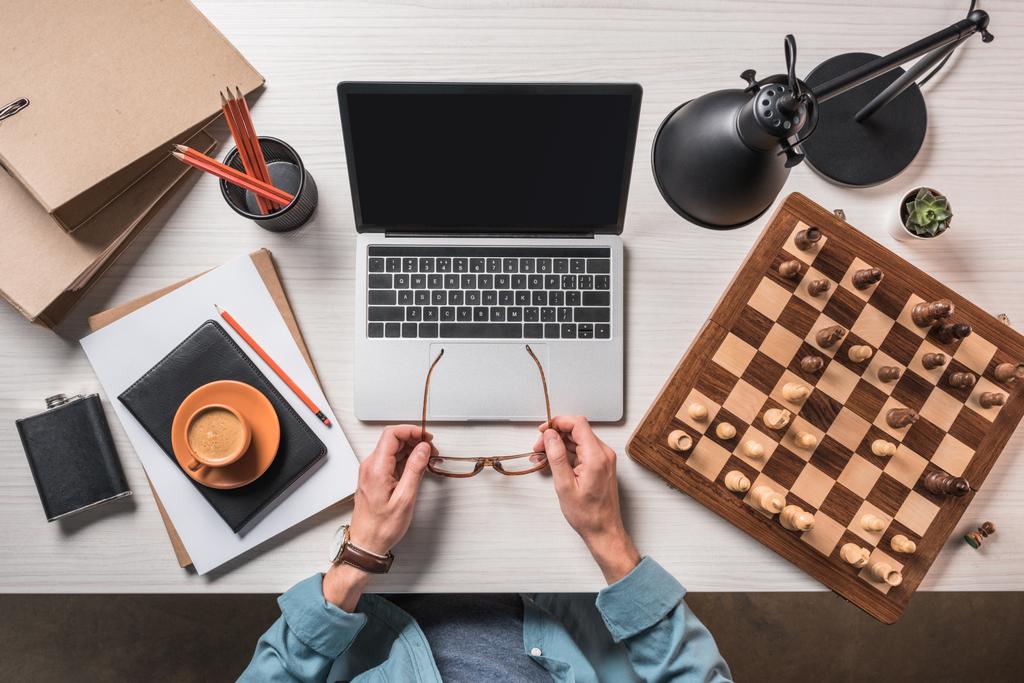 vista parcial del freelancer masculino con anteojos sentados en la mesa de trabajo con café, portátil y tablero de ajedrez
  - Foto, imagen