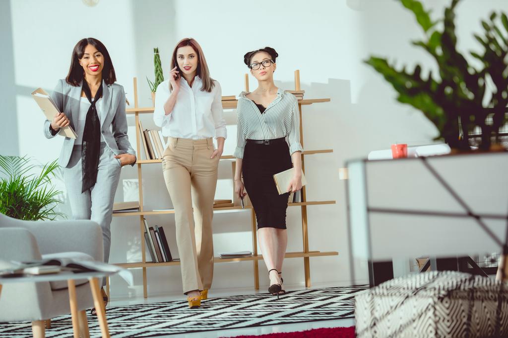 jóvenes mujeres de negocios multiétnicas sonrientes caminando juntas en la oficina
 - Foto, Imagen