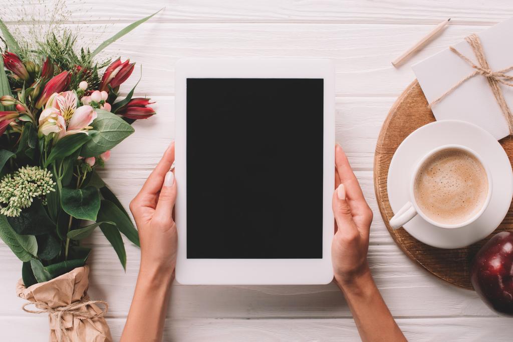 partial view of woman holding tablet with blank screen at surface with wrapped bouquet of flowers and cup of coffee - Photo, Image