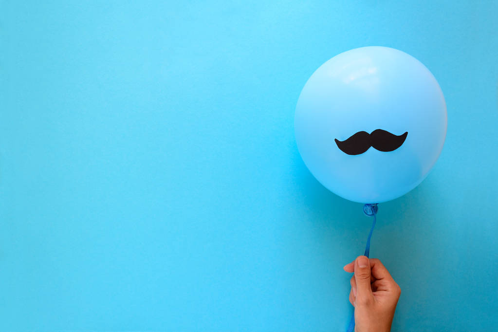Hand met blauwe ballon met een snor papier op blauw papier achtergrond. Knip stijl. Movember concept. Bovenaanzicht. Flat lag. Kopiëren van ruimte - Foto, afbeelding