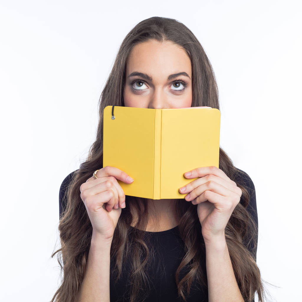 Porträt einer schönen jungen Frau mit langen Haaren, die ihren Mund mit einem gelben Notizbuch bedeckt und nach oben blickt. Studioaufnahme vor weißem Hintergrund.  - Foto, Bild