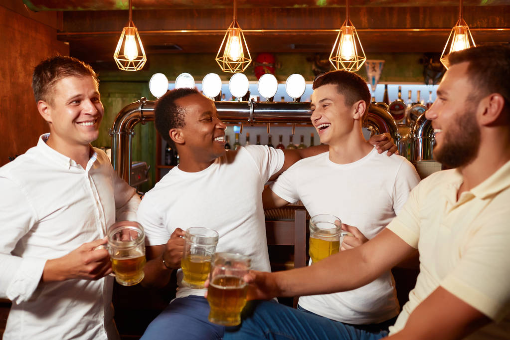 Компания из четырех красивых мужчин, держащих стакан пива, улыбающихся и обнимающих
. - Фото, изображение