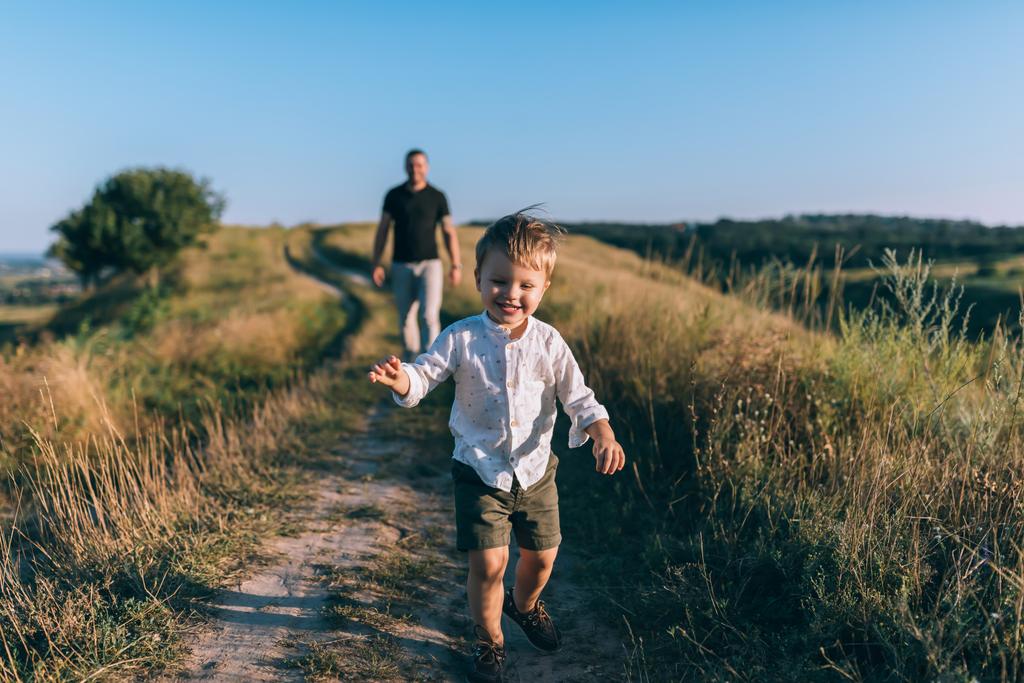 αξιολάτρευτο ευτυχισμένη μικρό αγόρι τρέχει σε αγροτική διαδρομή ενώ ο πατέρας το περπάτημα πίσω - Φωτογραφία, εικόνα