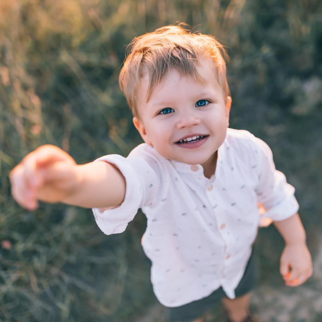 Blick aus der Vogelperspektive auf entzückend glückliches kleines Kind, das die Hand reicht und im Freien in die Kamera lächelt  - Foto, Bild