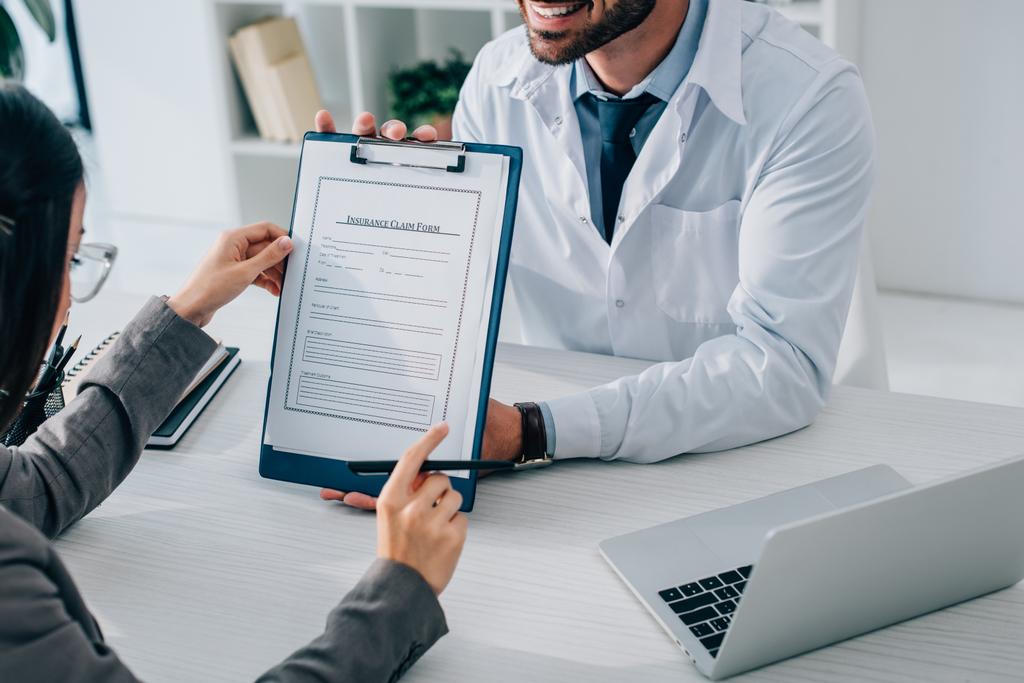 imagen recortada de médico sonriente mostrando formulario de reclamación de seguro al paciente en la clínica
 - Foto, Imagen