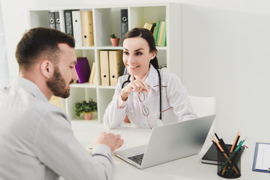 улыбающийся врач и человек обсуждают медицинскую страховку и глядя на экран ноутбука в клинике
 - Фото, изображение