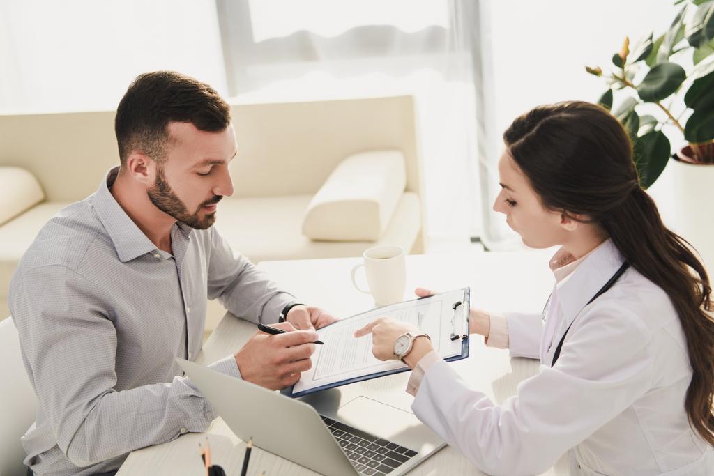 homme signant formulaire de demande d'assurance tandis que le médecin pointant vers elle dans le bureau avec ordinateur portable
 - Photo, image