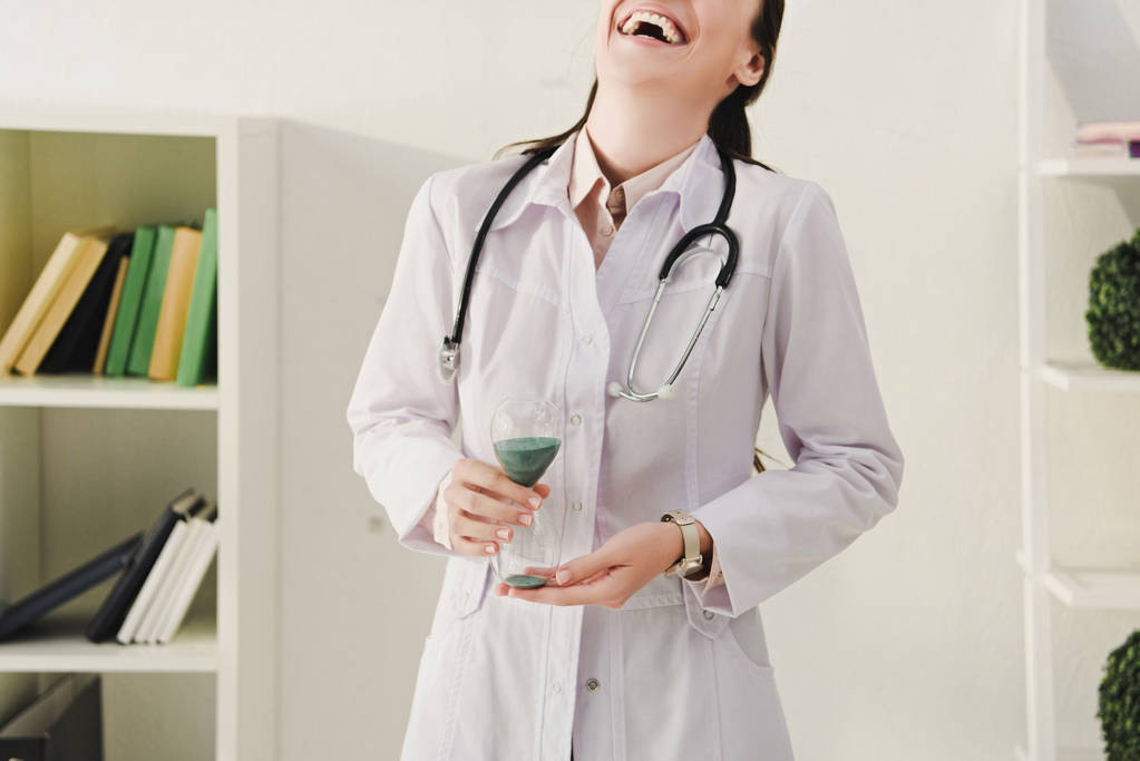cropped view of laughing doctor in white coat with stethoscope holding sand clock - Photo, Image