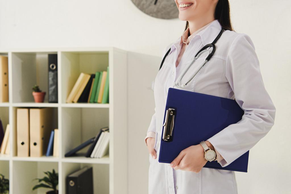 oříznutý pohled usmívající se doktor v bílém plášti s stetoskop drží schránky - Fotografie, Obrázek
