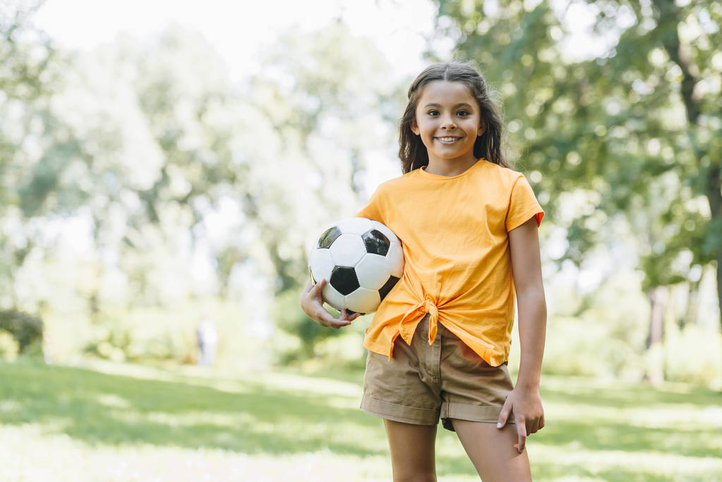 όμορφο ευτυχισμένο παιδί κρατώντας την μπάλα ποδοσφαίρου και να χαμογελά στη φωτογραφική μηχανή στο πάρκο - Φωτογραφία, εικόνα