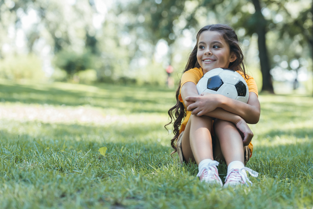 чарівна щаслива дитина тримає футбольний м'яч і дивиться в сторону, сидячи в парку
  - Фото, зображення