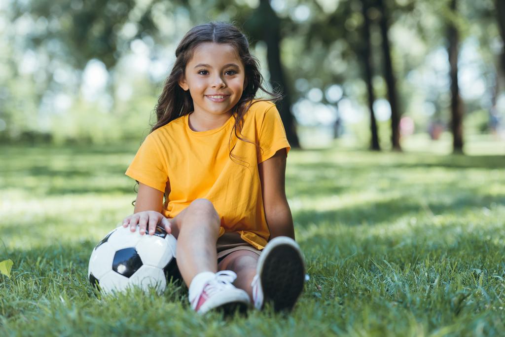 милий щасливий дитина сидить на траві з футбольним м'ячем і посміхається на камеру
 - Фото, зображення