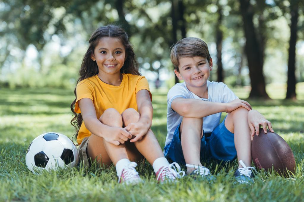 oturma ve kamera Parkta gülüp futbol ve ragbi topları ile şirin mutlu çocuk - Fotoğraf, Görsel
