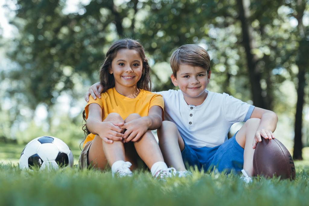 cute happy kids with soccer and rugby balls sitting on grass and smiling at camera - Photo, Image