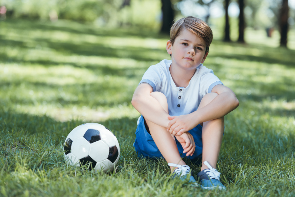 милий маленький хлопчик сидить на траві з футбольним м'ячем і дивиться на камеру в парку
  - Фото, зображення