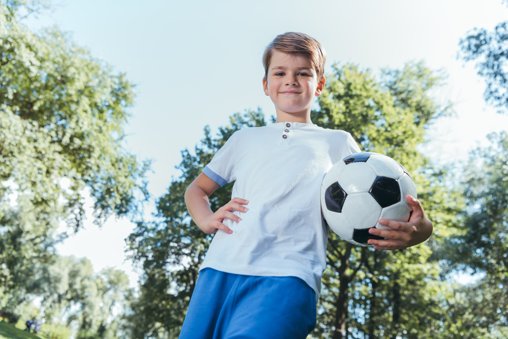χαμηλή γωνία θέα αγόρι κρατώντας την μπάλα ποδοσφαίρου και να χαμογελά στη φωτογραφική μηχανή στο πάρκο - Φωτογραφία, εικόνα
