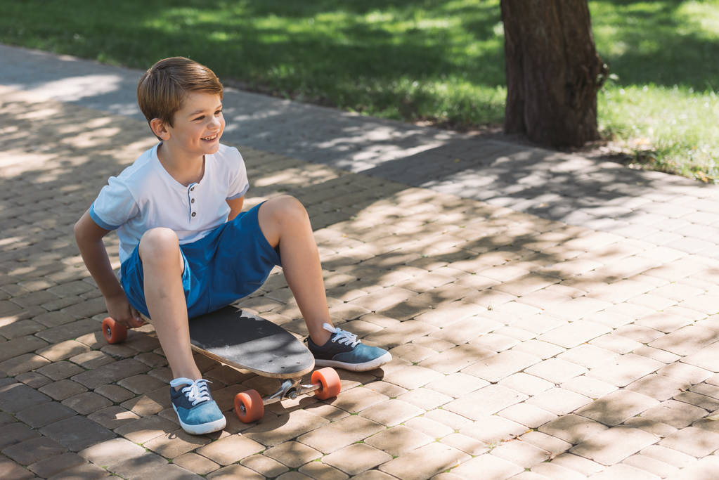 μεγάλη γωνία θέα χαμογελαστό αγόρι κάθεται στο skateboard και αναζητούν μακριά στο πάρκο - Φωτογραφία, εικόνα