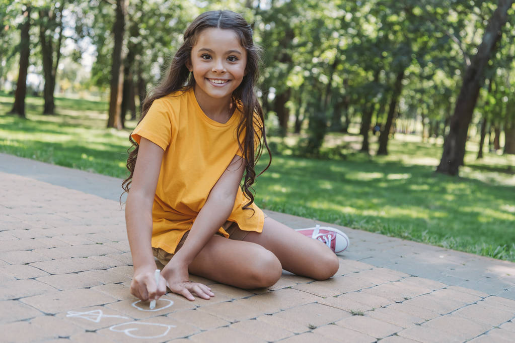 beautiful child drawing with chalk and smiling at camera in park - Photo, Image