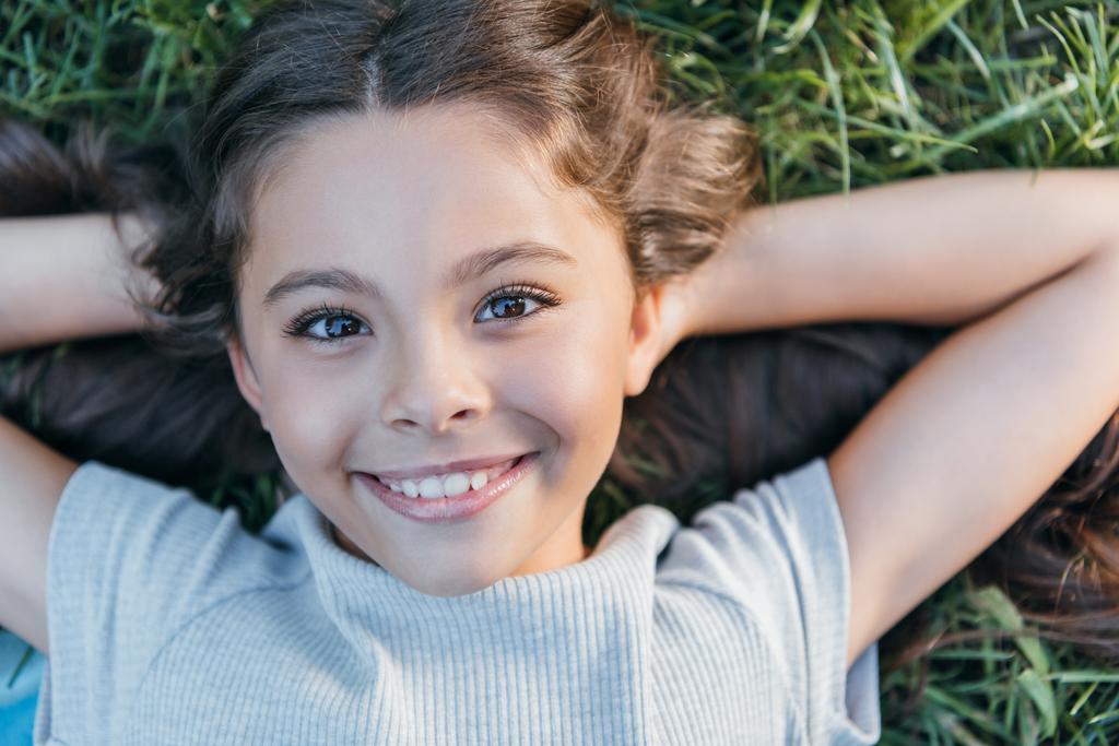 вид счастливого ребенка, лежащего с руками за головой на траве и улыбающегося в камеру
    - Фото, изображение