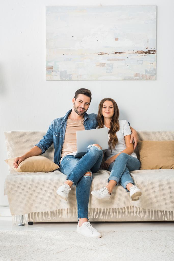 hymyilevä nuori pari istuu sohvalla kannettavan tietokoneen kanssa kotona
  - Valokuva, kuva