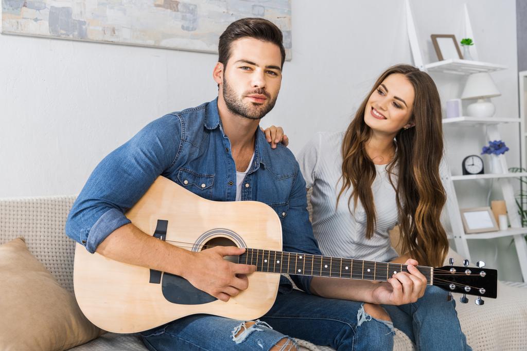 hombre guapo tocando la guitarra mientras su novia lo mira en el sofá en casa
 - Foto, imagen