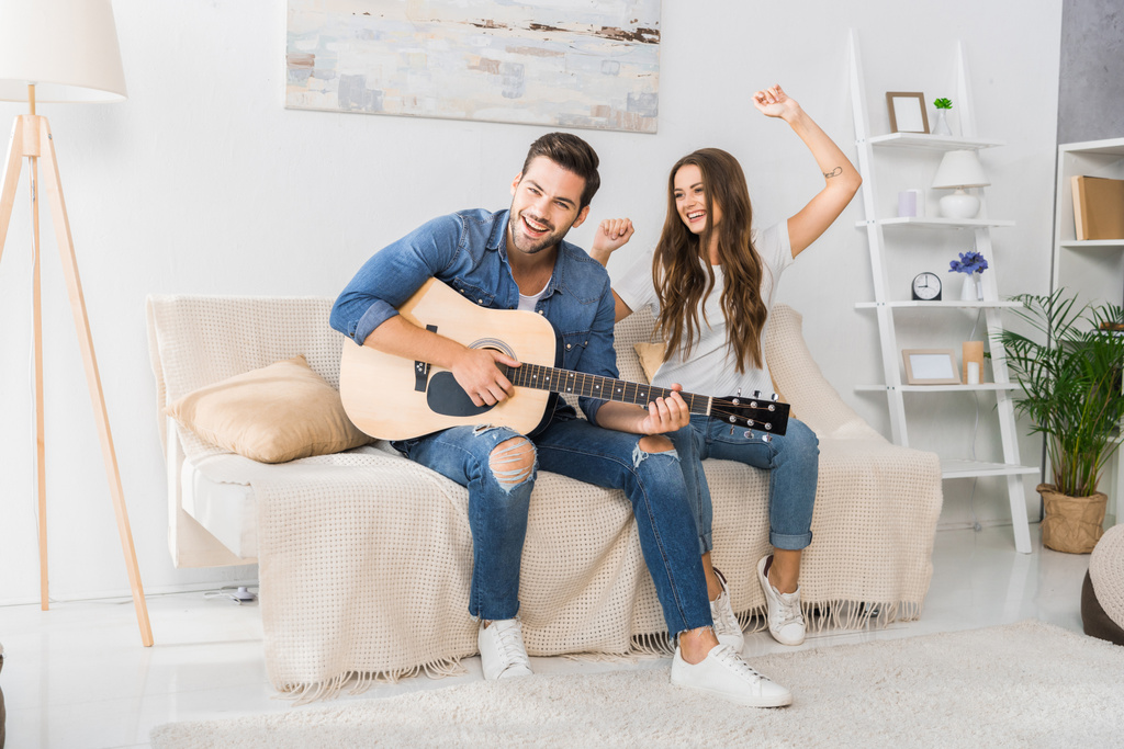 gülümseyen adam kız arkadaşı yakınındaki evde kanepede dans ederken akustik gitar çalmaya - Fotoğraf, Görsel