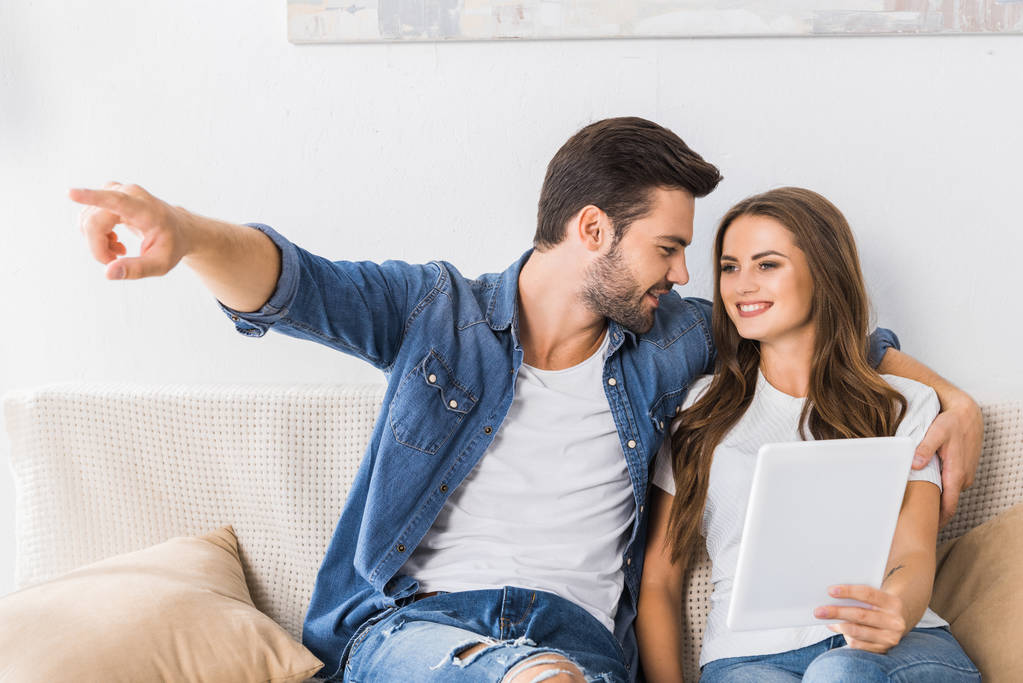 joven mirando a su novia con la tableta digital y señalando con el dedo a ella en el sofá en casa
  - Foto, Imagen