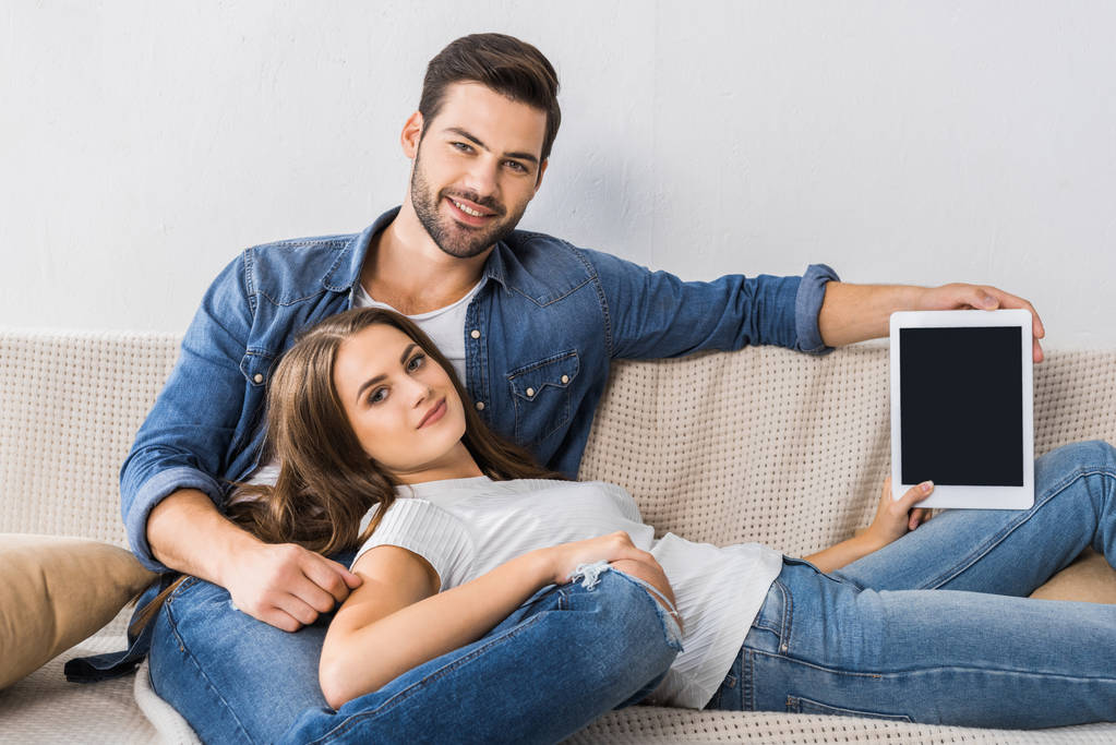 Lachende vrouw weergegeven: digitale tablet met leeg scherm terwijl haar vriendje in de buurt van zittend op Bank thuis  - Foto, afbeelding
