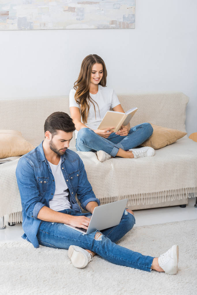 Selbstbewusster Mann mit Laptop sitzt auf dem Boden, während seine Freundin zu Hause auf der Couch Buch liest - Foto, Bild