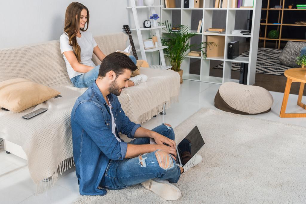 vista lateral del hombre sonriente usando el ordenador portátil y sentado en el suelo, mientras que su novia leer libro en el sofá en casa
 - Foto, Imagen