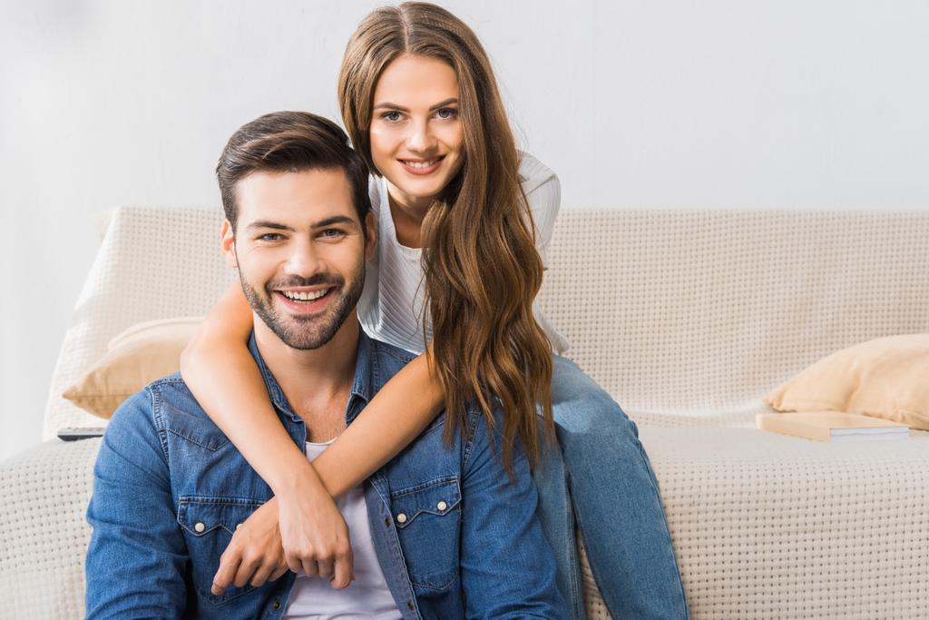 portrét mladého usmívající se pár na pohovce doma při pohledu na fotoaparát  - Fotografie, Obrázek