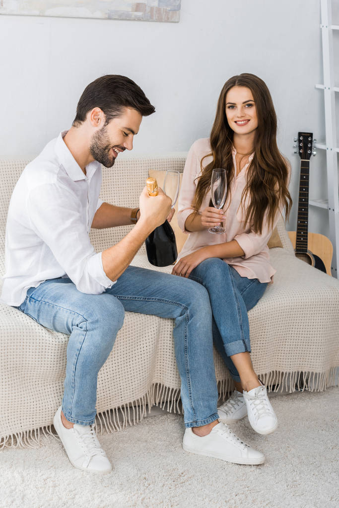 happy young couple celebrating with bottle of champagne and glasses on couch at home - Photo, Image