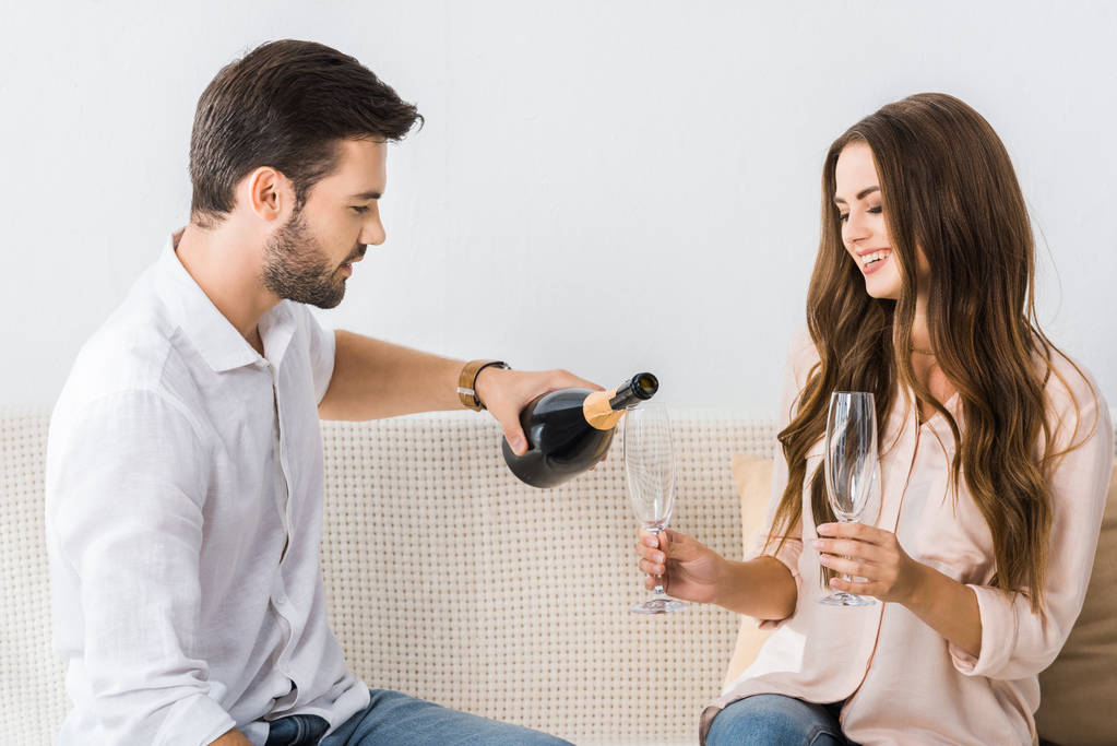 giovane uomo versando champagne in vetro mentre seduto sul divano con la ragazza a casa
  - Foto, immagini