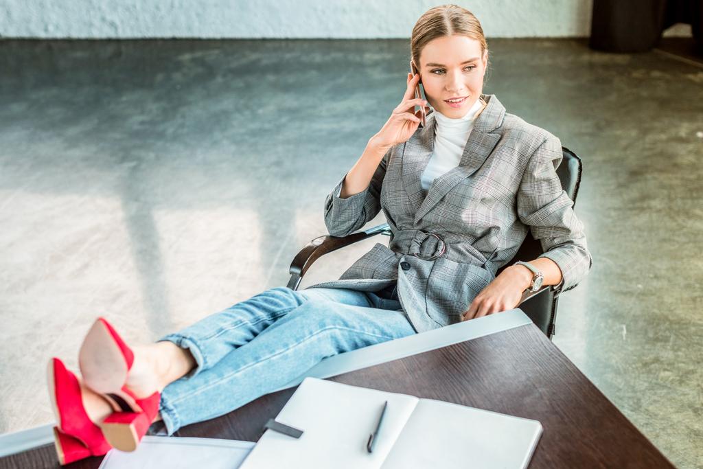 высокий угол обзора деловой женщины, сидящей с ногами на столе в офисе и разговаривающей по смартфону
 - Фото, изображение
