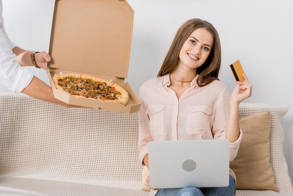 vista parcial del hombre con pizza en caja de papel y mujer sonriente con tarjeta de crédito y portátil en casa, concepto de compras en línea
 - Foto, imagen