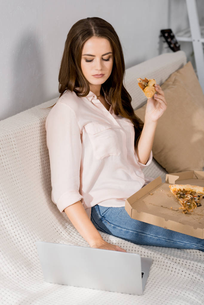 νεαρή γυναίκα με πίτσα χρησιμοποιώντας φορητό υπολογιστή ενώ ξεκουραζόταν στον καναπέ στο σπίτι - Φωτογραφία, εικόνα