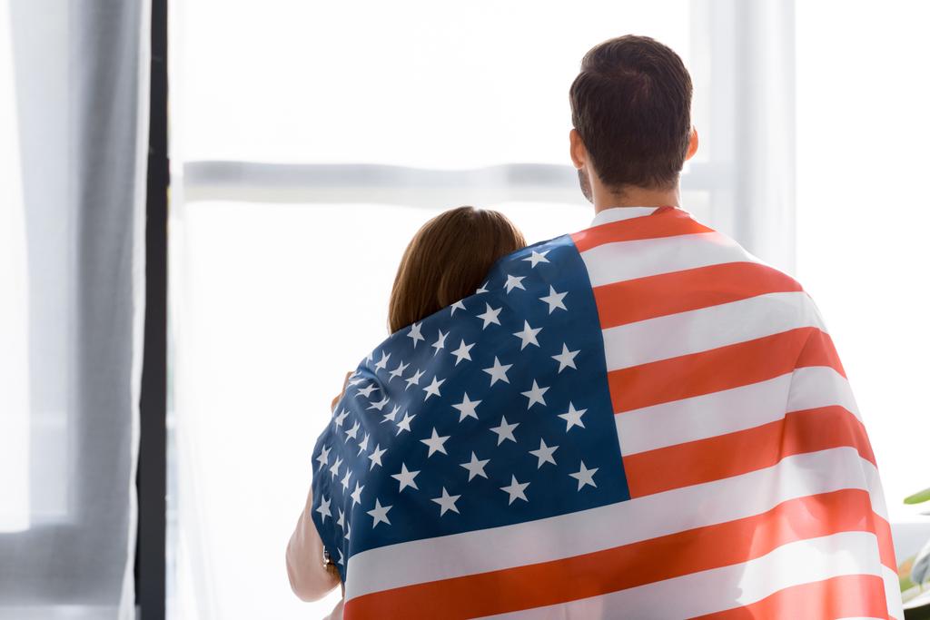 自宅の窓の外見ているアメリカの国旗で覆われてカップルの背面図 - 写真・画像