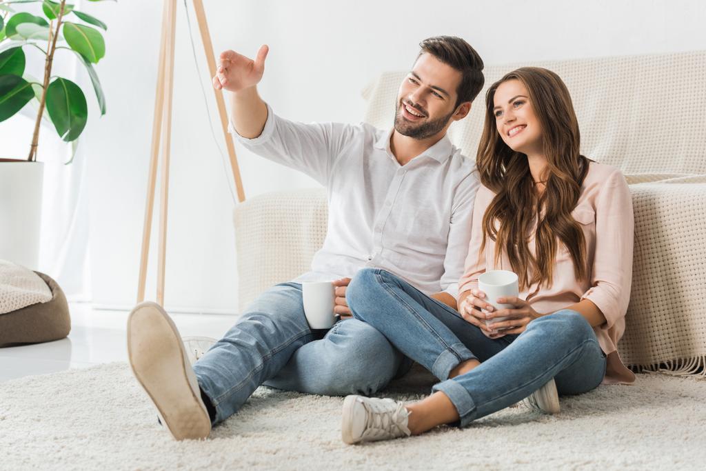 happy loving couple with cups of coffee sitting on floor and watching tv at home - Photo, Image
