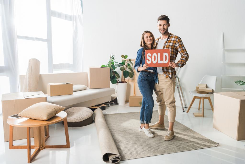 smiling couple holding sold red card at home with cardboard boxes - Photo, Image