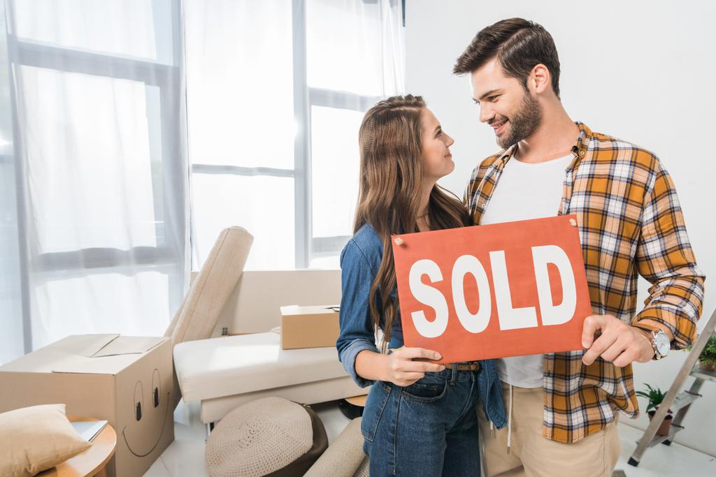 portrait of couple holding sold red card at home with cardboard boxes - Photo, Image