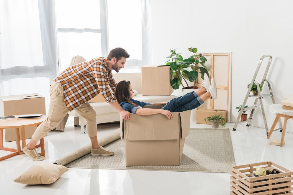 вид сбоку пары, веселящейся с картонной коробкой в новом доме, концепция переезда домой
 - Фото, изображение
