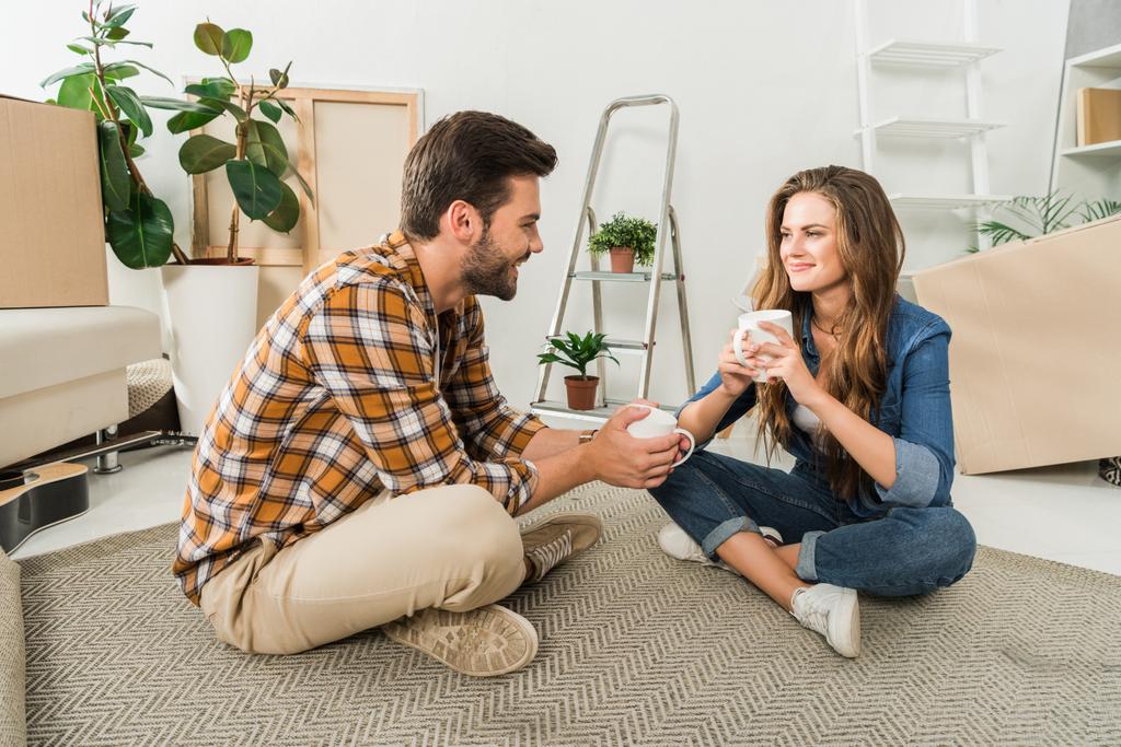 усміхнена пара з чашками кави сидить на підлозі в новому будинку, переміщуючи концепцію будинку
 - Фото, зображення