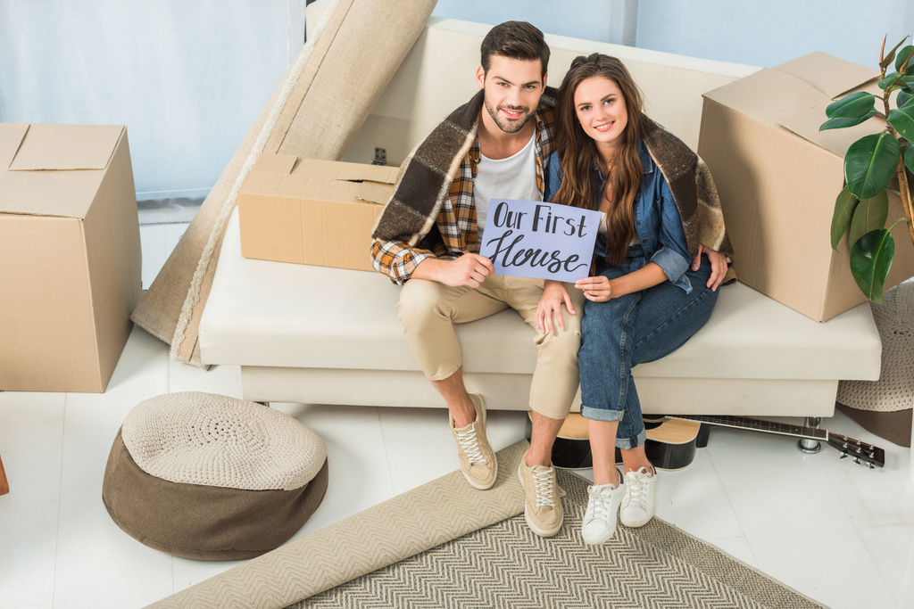 high angle view of young couple in blanket with our first house card sitting on sofa, moving home concept - Photo, Image